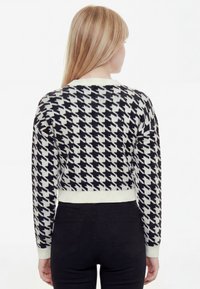 Žena s dlhými blond vlasmi, ktorá má na sebe sveter so vzorom čierno-bieleho psieho zubného vzoru a čierne nohavice, zobrazená zozadu.