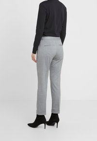 Persoon in lichtgrijze op maat gemaakte broek met omslag, zwart langemouwen top en zwarte hoge enkellaarsjes, staande tegen een effen achtergrond.
