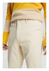 Pantaloni di cotone color crema con una texture liscia, dotati di chiusura frontale a bottoni e tasche laterali, abbinati a un maglione a righe gialle.