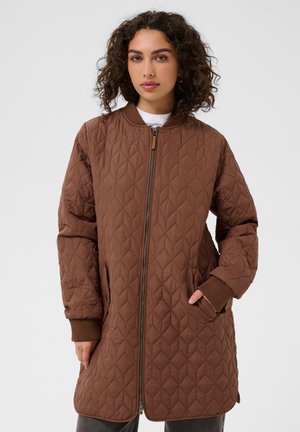 Femme aux cheveux bouclés portant une longue veste matelassée marron avec des poignets côtelés, une fermeture éclair sur le devant et des poches latérales, debout devant un fond uni.