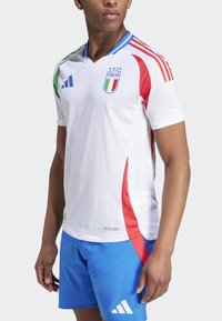Biely futbalový dres so zelenými, červenými a modrými akcentmi, s výstrihom do V, pruhmi Adidas a emblémom Talianska. Textúra je hladká a odvádzajúca vlhkosť.
