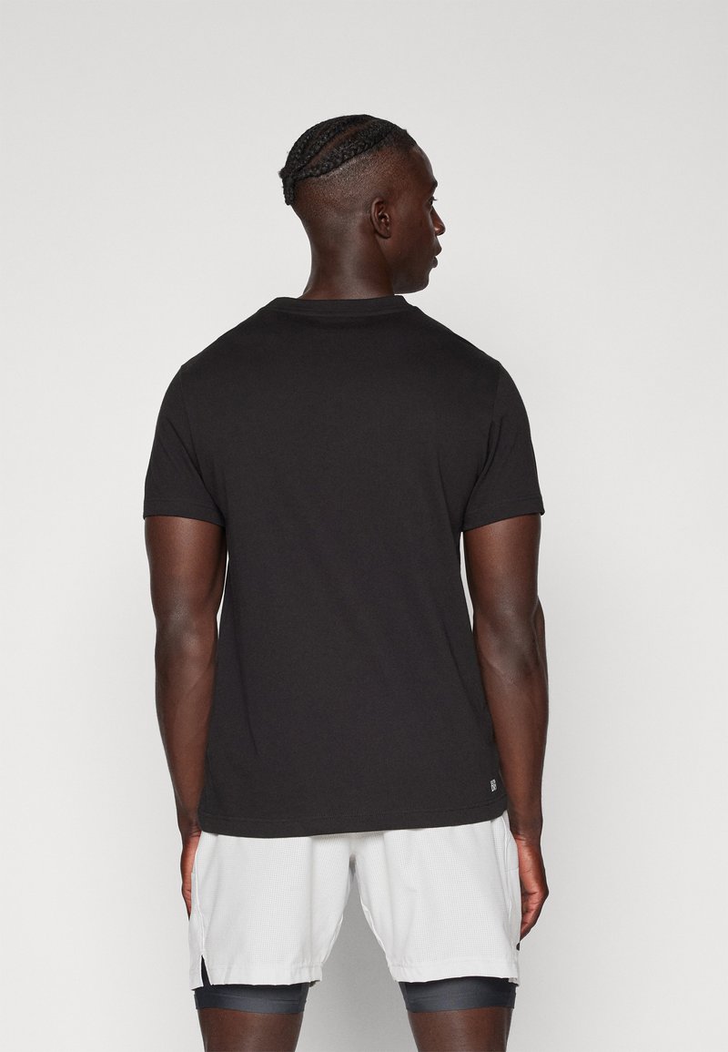 T-shirt en coton noir, à manches courtes, col rond. Présente un design épuré sans logos ni motifs visibles. Porté avec un short de couleur claire.