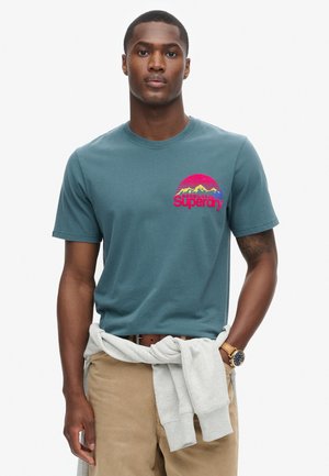 Teal bomulds t-shirt med et farvet grafisk design af bjerge og "Superdry" tekst. Modellen har beige bukser og en grå sweatshirt bundet omkring taljen.