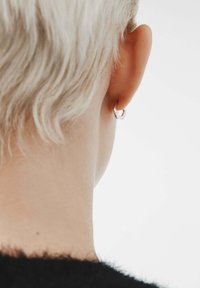 Pendiente de aro geométrico plateado en una oreja rubia clara, mostrando un diseño elegante con formas angulares y un estilo minimalista contra un fondo suave.