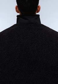 Giacca in pile nera con colletto alto, caratterizzata da un esterno texture morbidissimo e un design minimalista, focalizzata su calore e comfort.