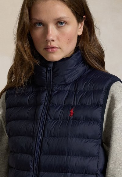 Jonge vrouw met lang bruin haar die een marineblauw doorgestikt vest draagt met rood geborduurd logo over een grijze longsleeve.