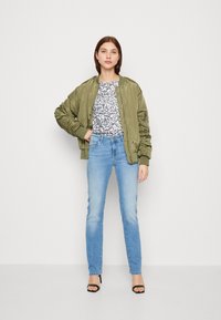 Chaqueta bomber verde oliva, top con estampado de leopardo gris, jeans claros y sandalias de tacón negro; modelo posa frente a un fondo neutro.