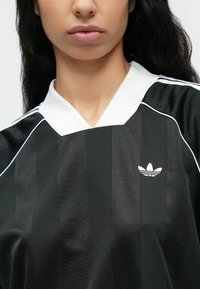 Čierny atletický top s vertikálnymi pruhmi, bielym golierom a vyšívaným bielym logom Adidas na hrudi. Štýlová, textúrovaná látka.