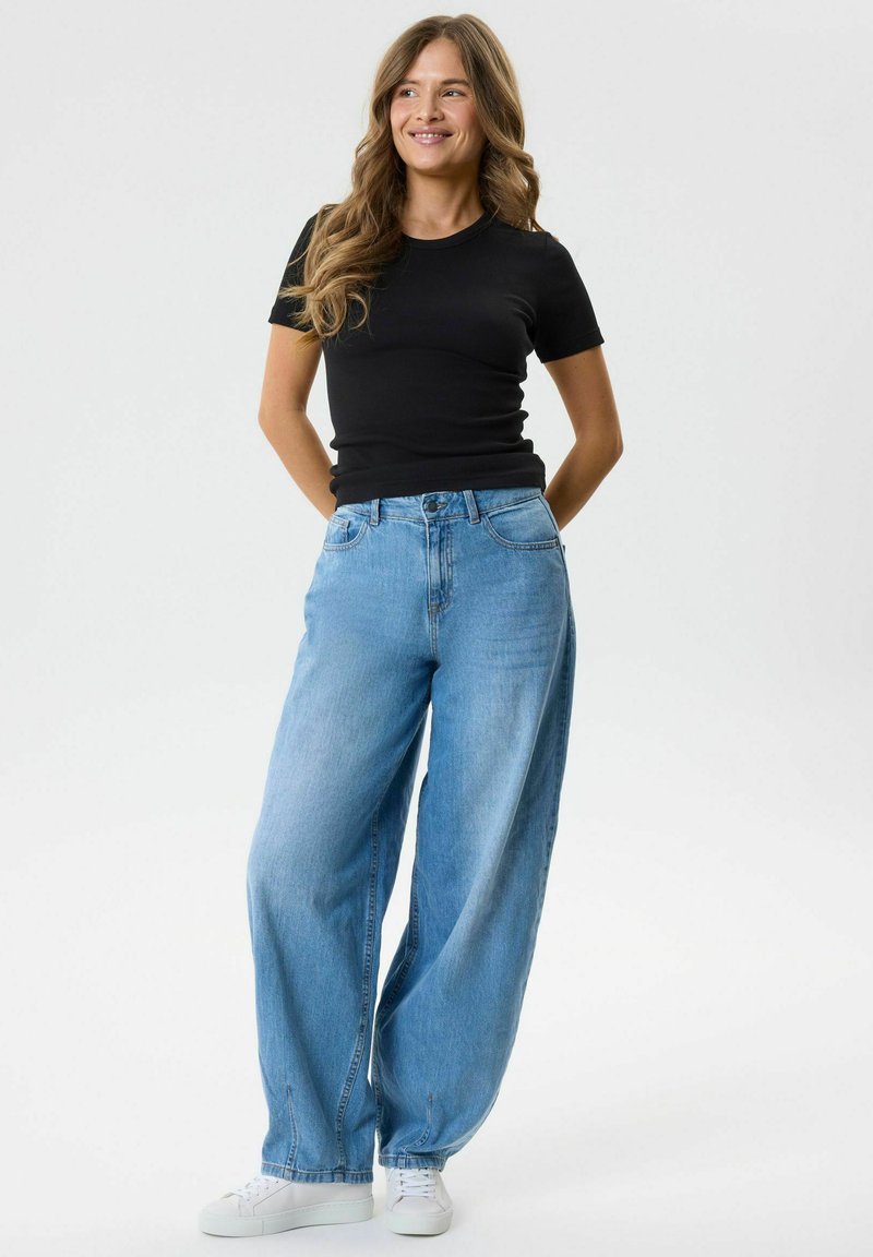 Femme aux longs cheveux ondulés porte un haut noir à manches courtes, un jean bleu clair à jambes larges et des baskets blanches, debout les mains dans le dos.