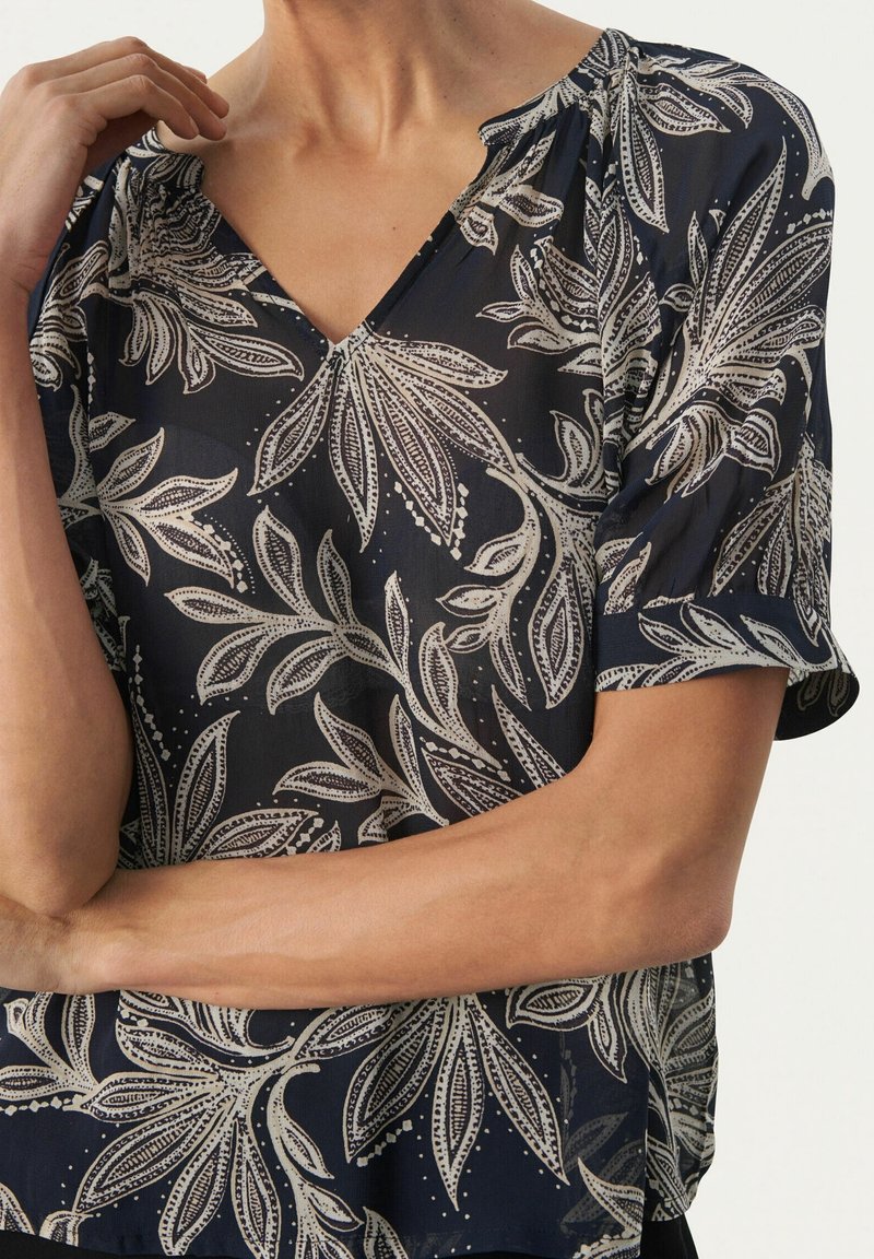Blusa navy con maniche corte, scollo a V e stampa floreale in crema con dettagli intricati. Tessuto leggero e trasparente con una vestibilità morbida.