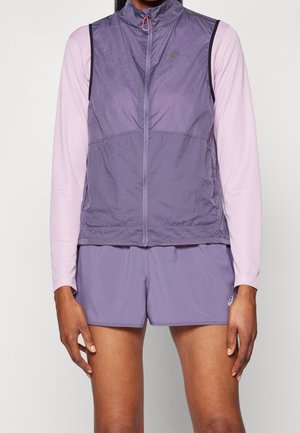 Chaleco ligero sin mangas color púrpura con cremallera frontal, tejido texturizado y ribete negro. Combinado con una camiseta de manga larga rosa y pantalones cortos a juego.