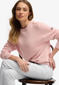 Pull rose clair en tricot avec un col rond et des poignets côtelés, orné d'un petit logo sur la manche. Associé à un pantalon blanc.