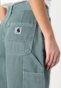 Lyse grønne bukser lavet af denim, med en baglomme med et rektangulært logo-label og sidelommer. Simpelt, funktionelt design.