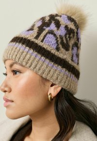 Beige Strickmütze mit lila und braunen Mustern. Verfügt über einen gerippten Bund und einen flauschigen Pompon oben. Weicher Wollstoff.