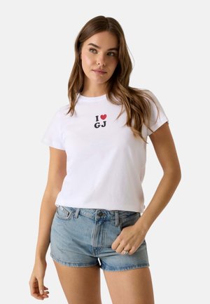 Donna con capelli castani lunghi che indossa una maglietta bianca con la scritta "I [cuore] GJ" e pantaloncini di jeans azzurri, in posa davanti a uno sfondo semplice.