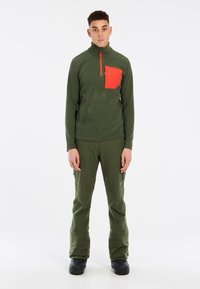 Olijfgroen thermoshirt met een rode ritszak op de borst en een kwart-rits kraag; gecombineerd met bijpassende olijfgroene broek en zwarte schoenen.