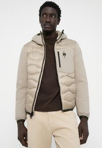 Beige pufferjack met een gestikte patroon en een capuchon. Voorzien van een ritszak, contrasterende bruine binnenvoering en zwarte ritsaccenten.