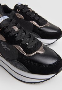 Černé tenisky s kombinací semiše a kůže, kovovými akcenty, texturovanými vrstvami a černými tkaničkami na bílých a modrých pruhovaných podrážkách.