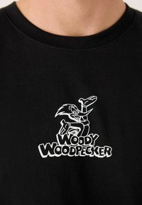 Čierne bavlnené tričko s bielou karikatúrou Woodyho Woodpeckera a textom "WOODY WOODPECKER" nižšie. Okrúhly výstrih navrhnutý pre pohodlie.