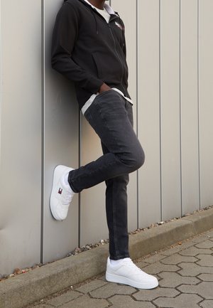 Person trägt schwarzen Hoodie, dunkle Jeans und weiße Sneaker, lehnt mit einem Fuß an einer grauen Metallwand.