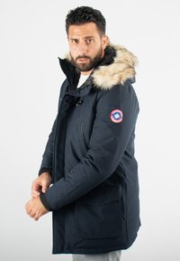 Parka navy avec capuche en fausse fourrure, fermeture éclair à l'avant et deux poches latérales. Les poignets côtelés ajoutent de la texture. Présente un badge logo sur la manche.