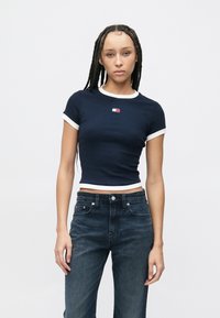 Námornícky modrý krátky top s bielym lemovaním, rebrovanou textúrou a malým logom na hrudi. Kombinovaný s tmavomodrými džínsami v klasickom strihu.