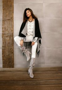 Grijze en zwarte poncho met franjes, witte gebreide trui, witte broek en grijze kniehoge laarzen met een blokhak, tegen een neutrale achtergrond.