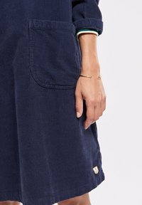 Robe en velours côtelé marine avec une poche, des manches retroussées et un petit logo d'ancre à l'ourlet. Texture douce avec un motif à côtes verticales.