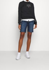 Sudadera negra con el logo "TOMMY JEANS", camisa de collar blanca debajo, pantalones cortos de mezclilla azules y zapatillas blancas. Atuendo simple y casual.