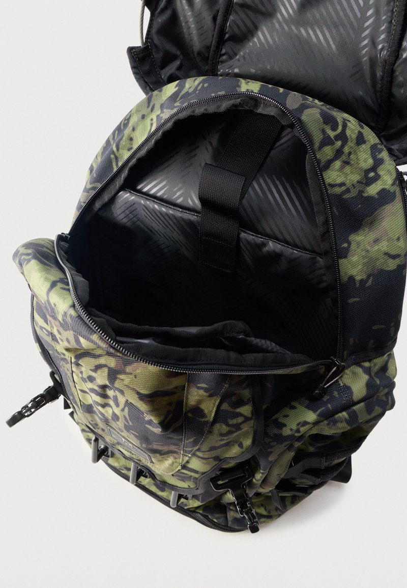 Grøn og sort camouflage-rygsæk åben for at vise det rummelige indre med polstret rum og sort strop indeni.