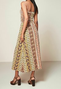 Robe maxi bustier à motifs multiples en jaune, rouge et marron, avec un corsage froncé et une jupe évasée. Portée avec des sandales à talons hauts.