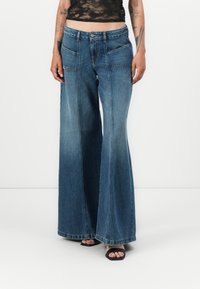 Jeans en denim à jambe large de couleur bleu foncé, avec un design classique à cinq poches, une coupe décontractée et un ourlet brut. Associés à des sandales à talons noires.