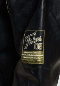 Ærmet på den sorte læderjakke prydes af et fremtrædende hvidt og guld Fubu-logo, med tekst der beskriver brandets engagement i kvalitet.