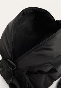 Sac à dos noir en nylon avec fermeture éclair principale partiellement ouverte, montrant un intérieur spacieux et vide ainsi qu'une poche avant avec boutons-pression et sangle réglable.