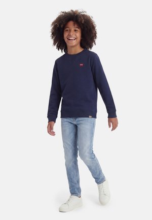 Naeratav laps lokkis juustega, kandes mereväe sinist dressipluusi, helesiniseid teksaseid ja valgeid tossusid ning kõndides ühe jalga tõstetud.