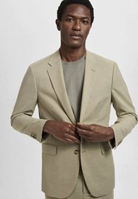 Lichtbeige blazer met een enkele knoopsluiting, twee voorkezen en een gestructureerde stof. Gedragen over een grijze ronde hals trui.