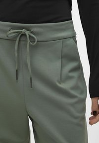 Lichtgroene sweatpants met een trekkoord in de taille, voorzien van zijzakken en een gladde textuur, gedeeltelijk zichtbaar tegen een zwarte top.