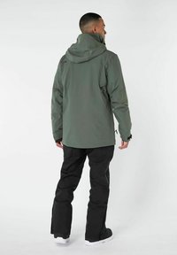 Giacca impermeabile verde con cappuccio, pantaloni neri e calzature bianche. Caratteristiche includono una vestibilità rilassata e tasche laterali con zip.