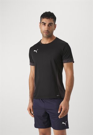 Schwarzes Sport-T-Shirt mit kontrastierenden lila Ärmeln, mit einem weißen Logo auf der Brust; getragen mit dunklen Shorts.