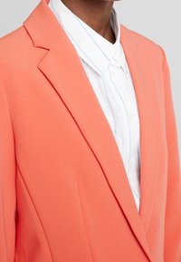 Koralowa marynarka o dopasowanym kroju, z wyciętymi klapami. W zestawie z białą koszulą z jasnoniebieskimi paskami. Gładka faktura.