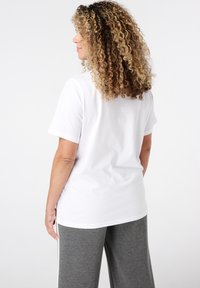 Femme aux cheveux bouclés portant un t-shirt blanc uni et un pantalon gris, se tenant dans une pose détendue face à gauche sur un fond blanc.