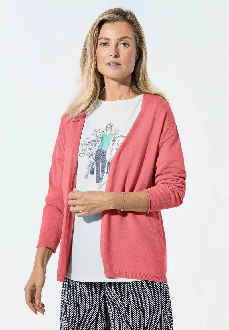Korallefarbener Cardigan mit langen Ärmeln und offener Vorderseite, über einem weißen T-Shirt mit Grafikumdruck, das eine Modeillustration zeigt, kombiniert mit schwarz-weißen gemusterten Hosen.