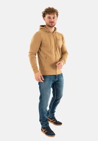Felpa beige con zip e tasca frontale, abbinata a jeans blu e sneakers nere con dettagli marroni. Tessuto liscio, vestibilità casual.