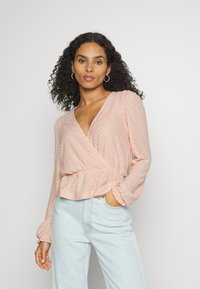 Blusa envolvente rosa claro com tecido texturizado, decote em V profundo, mangas bufantes e cintura franzida, combinada com jeans azul claro.