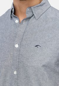 Camisa de botones gris hecha de una tela texturada, con un pequeño logo en navy en el pecho y botones blancos a lo largo de la tapeta.