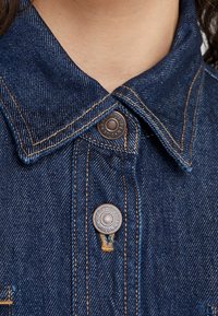 Denimjacke in dunkler blauer Farbe, mit spitzem Kragen, zwei Metallknöpfen und kontrastierenden gelben Nähten.