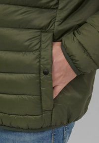 Olivgrüne Daunenjacke mit horizontalen Nähten, Seitentasche und elastischen Bündchen. Das Material wirkt glatt und leicht glänzend.