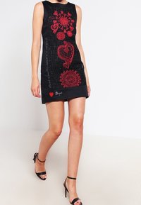 Kvinna som bär en ärmlös svart mini-klänning med röda blommor och hjärtmönster, kombinerat med svarta högklackade sandaler med ankelrem, som går.