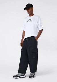 Weißes kurzärmeliges T-Shirt mit schwarzem Logo, kombiniert mit lockeren schwarzen Cargo-Hosen mit Taschen, dazu graue und schwarze Sneakers und eine schwarze Mütze.