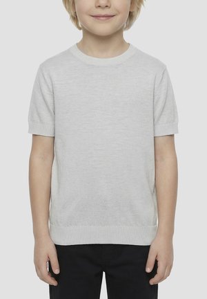 Enfant à la peau claire et aux cheveux blonds portant un pull léger gris clair uni à manches courtes et un pantalon noir, debout devant un fond neutre.